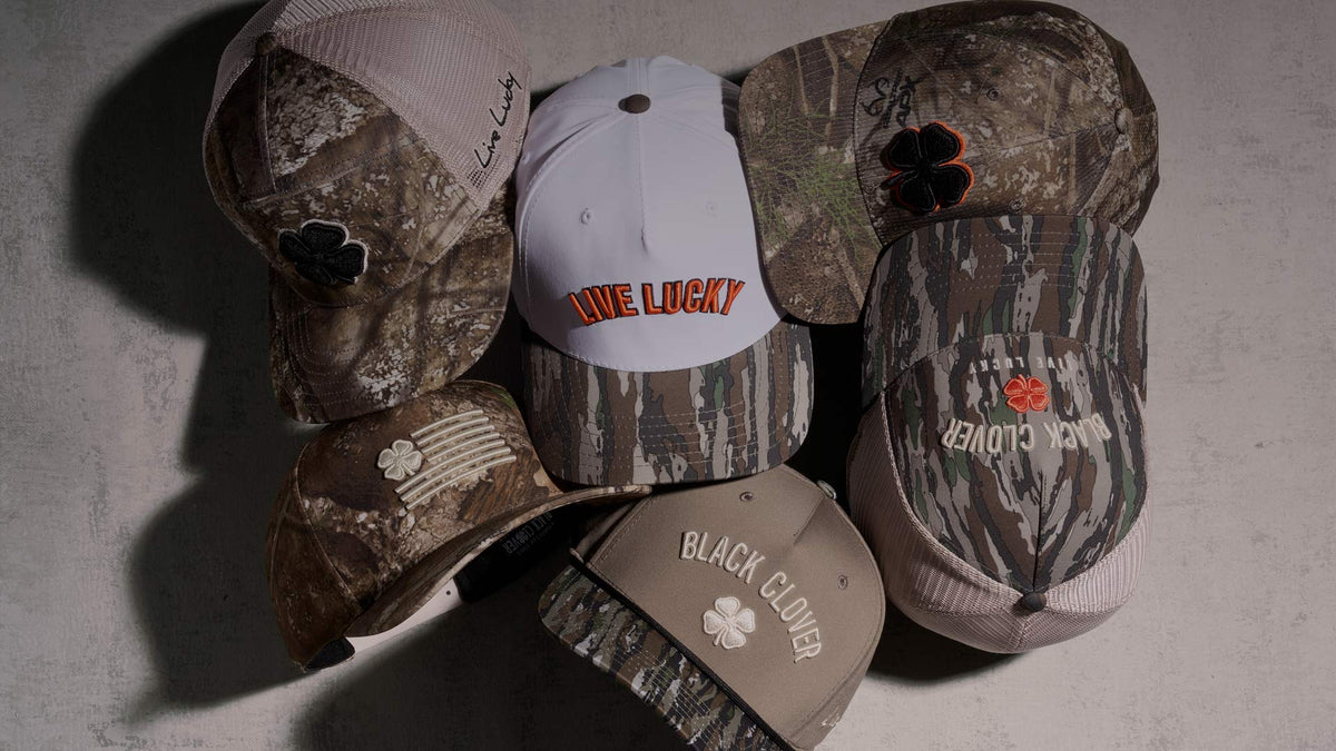 Six baseball caps in various camouflage and solid color designs are arranged in a fan-like pile on a textured gray background. The caps feature different patterns including woodland camo, coyote brown camo, and solid beige/white, with various embroidered logos such as 'Black Clover', 'Live Lucky', and a clover patch.
